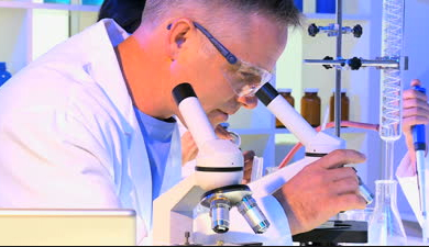Picture of a Lab Tech sitting down and looking into a microscope with safey glasses on.