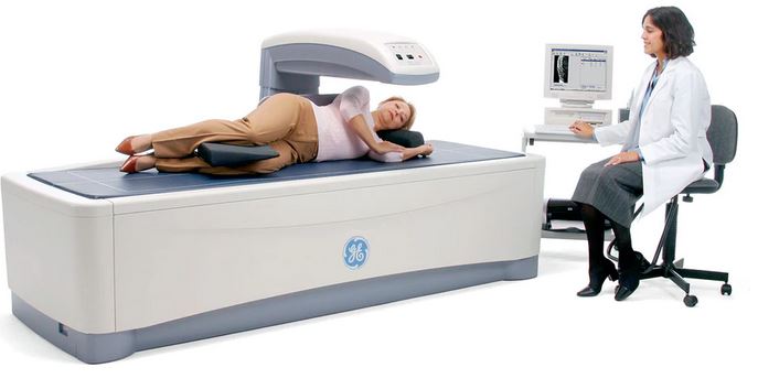 Picture of a female lying down on a bone density scanning machine and there is a female X-Ray Tech sitting in a rolling chair next to a screen.